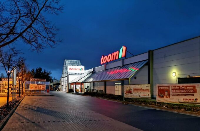 Toom Baumarkt bei Dämmerung, dessen beleuchtetes Logo gut sichtbar auf dem Dach des Gebäudes zu sehen ist. Der Eingangsbereich ist ebenfalls beleuchtet, während der Parkplatz leer und ruhig wirkt. Es sind Werbebanner an der Außenwand des Gebäudes angebracht, die auf verschiedene Angebote und Dienstleistungen hinweisen.